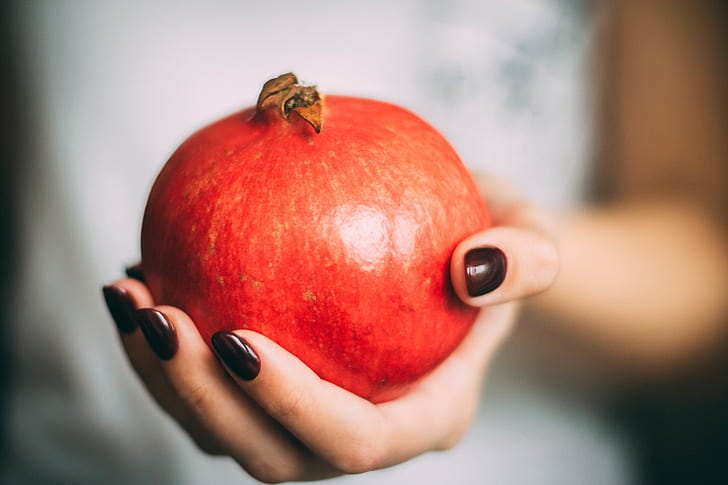 Picture shows a pomegranate in a pale-skinned hand with dark brown nail polish - the background is blurred, appearing to show the torso and arm of a person wearing white thrusting the fruit at the viewer