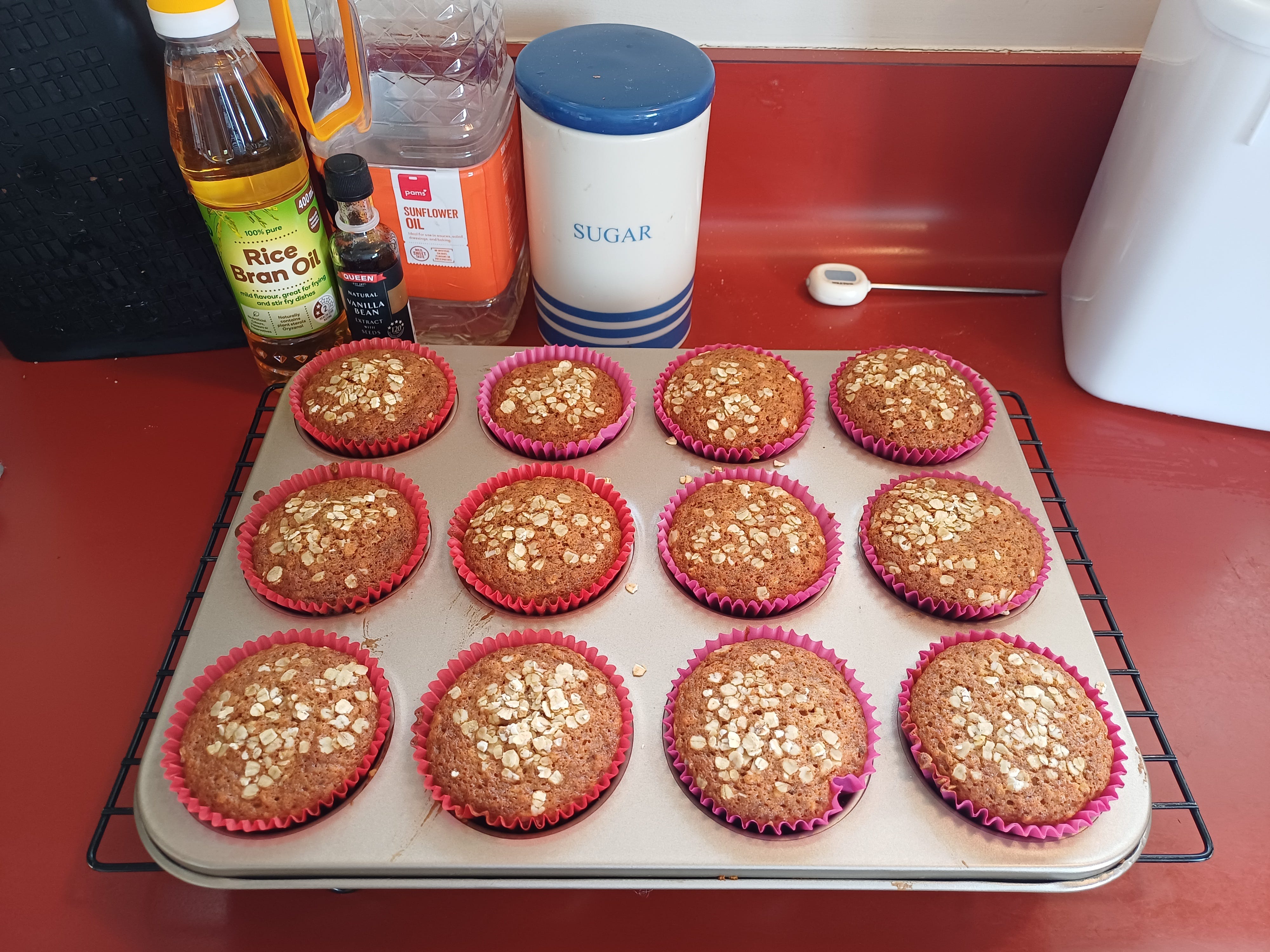 A tray of delightfully brown muffins in a tray, sitting on a rack set on a red bench. In the background are some of the ingredients, looking suspiciously neat, and also a blue and white cannister labelled "SUGAR". This is a lie. That's where I keep my salt.